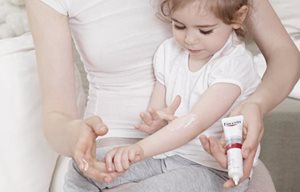Niña pequeña, sentada en el regazo de su madre, aplicándose Eucerin AtopiControl Crema Forte en la cara interna del brazo.